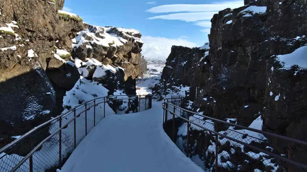 A stunning view of Almannagjá Gorge in Iceland, featuring a snowy path leading through rugged volcanic rock walls. The path is lined with a metal railing for safety, and the rocks on either side are dark and imposing, partially covered in snow. The scene is illuminated by a clear blue sky, creating a stark contrast between the dark rocks and the bright snow. In the distance, a view of the landscape unfolds, showing more of the gorge's rocky terrain. This image captures the raw and breathtaking beauty of Iceland's geological formations, highlighting the dramatic landscapes found in the country.