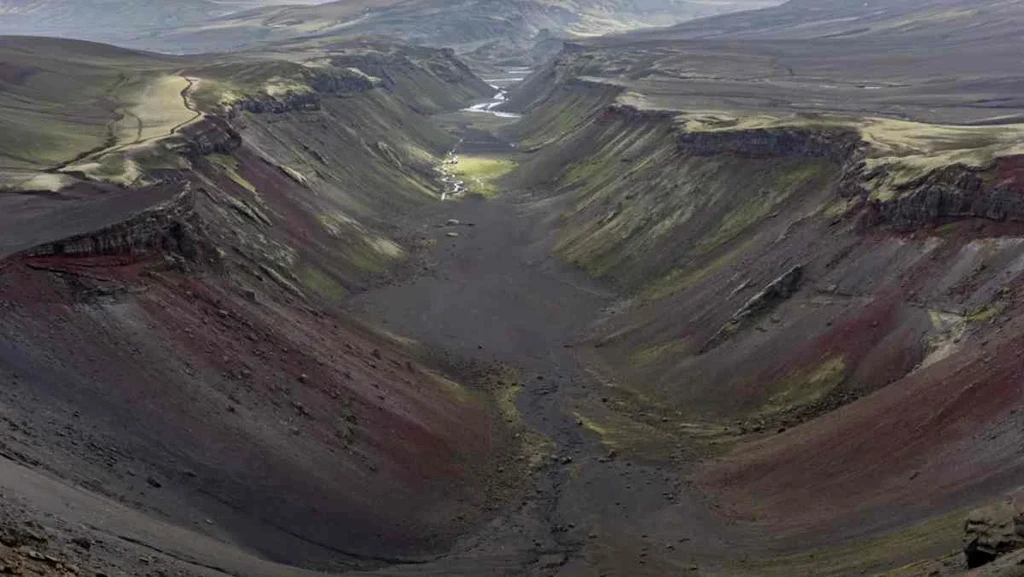 A view of Eldgjá Canyon in Iceland, showcasing its expansive, barren landscape with steep walls and a narrow river running through the center. The canyon, formed by volcanic activity, displays shades of red and brown, highlighting its geological features. The scene captures the rugged and dramatic nature of Iceland's volcanic terrain.