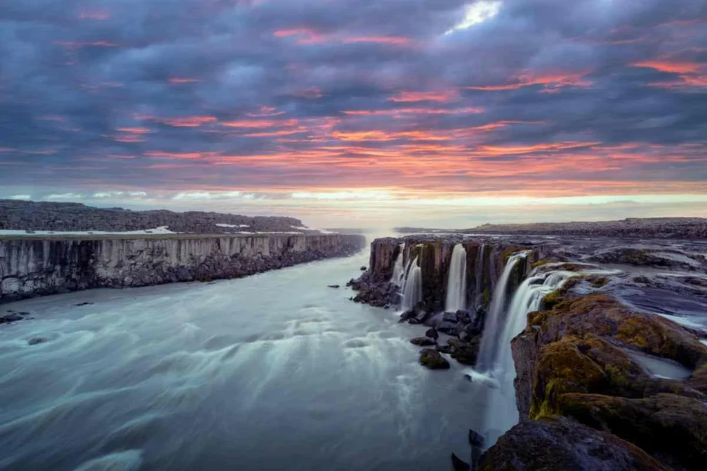 A stunning view of Jokulsargljufur Canyon in Iceland at dusk, with vibrant colors of pink and purple in the sky. The scene features powerful waterfalls cascading over rocky cliffs into a fast-flowing river below. The canyon walls are rugged and covered in dark basalt formations, contrasting with the smooth, flowing water. The landscape captures the raw beauty and dramatic atmosphere of this natural wonder, highlighting the unique geological features formed by glacial activity and volcanic processes. The peaceful yet powerful scenery offers a breathtaking glimpse into the majestic landscapes of Iceland.