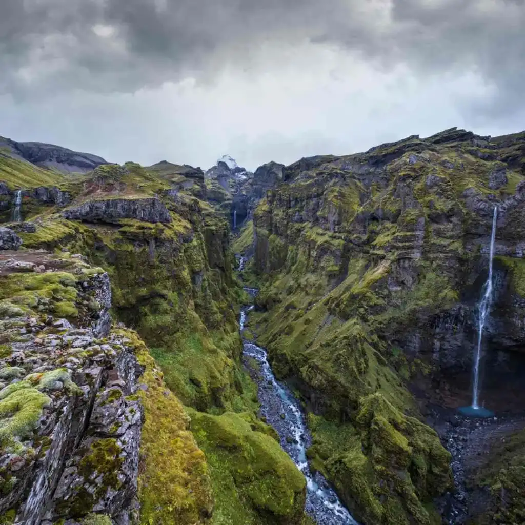 A breathtaking view of Múlagljúfur Canyon in Iceland, showcasing its dramatic landscape. The canyon is flanked by steep, rugged cliffs covered in lush green moss and vegetation. A narrow river winds its way through the canyon, flanked by towering rock walls on either side. Several small waterfalls cascade down the cliffs, adding to the serene and mystical atmosphere of the scene. The sky above is overcast, casting a moody light over the landscape, enhancing the natural beauty and untouched wilderness of the area. This image captures the awe-inspiring scenery of one of Iceland's hidden gems.