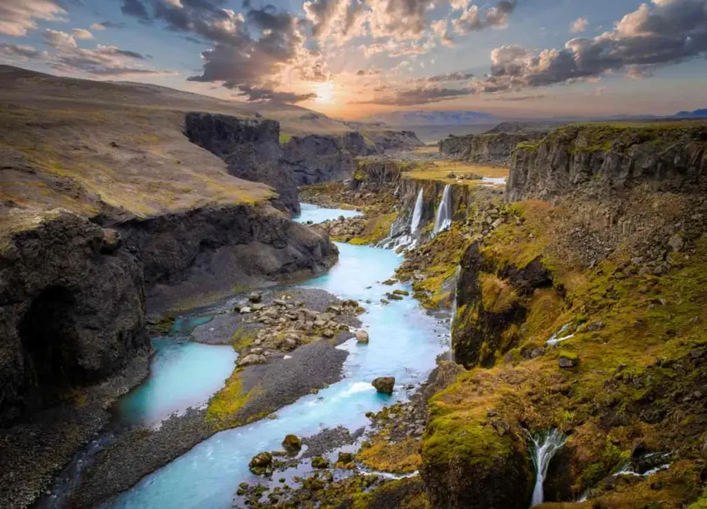 A breathtaking view of Sigoldugljufur Canyon in Iceland, showcasing a series of waterfalls cascading into a turquoise river winding through the rugged landscape. The canyon is surrounded by steep cliffs covered in green moss and rocky terrain. The scene is bathed in warm, golden light from the setting sun, with clouds scattered across the sky. The vibrant colors of the water and the lush greenery contrast beautifully with the dark, volcanic rock formations, creating a stunning natural panorama.