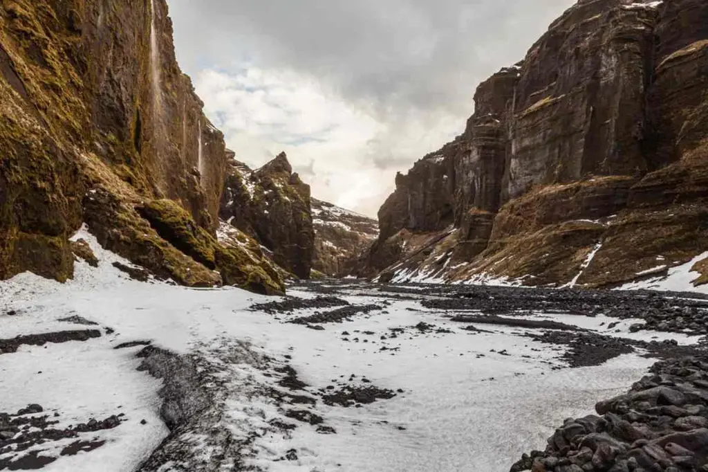 A dramatic view of Stakkholtsgja Canyon in Iceland, showcasing towering rock walls lined with patches of moss and snow. The canyon floor is covered in a thin layer of snow, with dark volcanic rocks scattered throughout. The rugged, steep cliffs rise high on either side, creating a narrow passage that stretches into the distance. The sky above is overcast, casting a soft light that accentuates the textures and colors of the rocks. This image captures the stark and rugged beauty of Iceland's natural landscapes, highlighting the raw and untouched nature of the canyon.