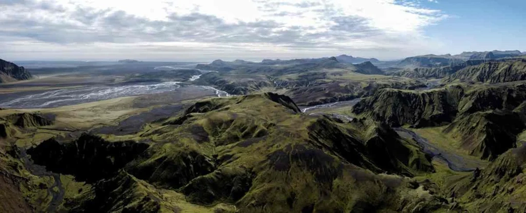 A stunning aerial view of Þakgil Canyon in Iceland, displaying a vast expanse of rugged terrain. The landscape is characterized by rolling green hills and deep valleys, with a network of rivers and streams meandering through the area. The terrain is covered in patches of vibrant moss and other vegetation, adding a touch of color to the otherwise earthy tones. In the distance, the landscape stretches out towards the horizon, with mountains and vast plains visible under a partly cloudy sky. The image captures the raw and untamed beauty of Iceland's wilderness, showcasing the unique geological formations and natural beauty of Þakgil Canyon.