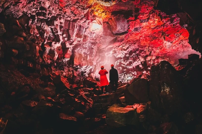 Two people explore the vibrant interior of a lava cave, illuminated by red and orange lights reflecting off the rocky walls.