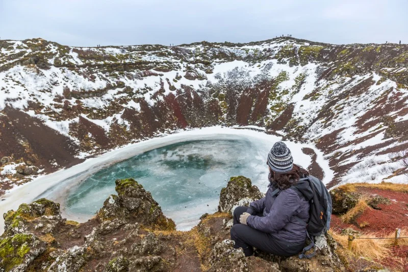 A person in winter clothing sits on a rocky edge overlooking the frozen Kerid Crater Lake, surrounded by snow-covered volcanic landscape.