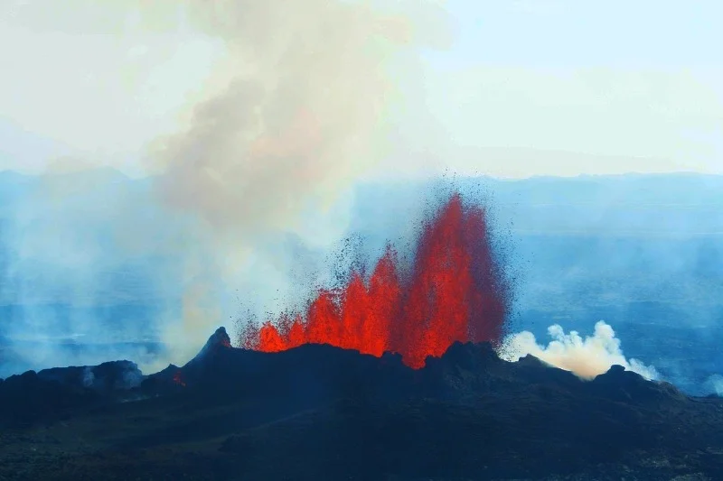 Dramatic eruption of a volcano with bright red lava fountains and thick smoke against a hazy sky, showcasing the power of nature.