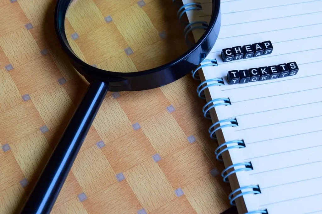 Magnifying glass on a wooden surface next to a notebook with the words 'cheap tickets' spelled out, symbolizing the search for affordable travel options