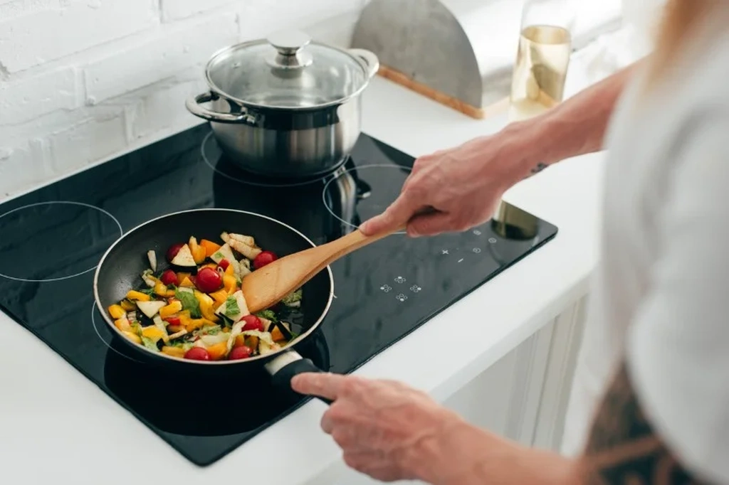 Person cooking a colorful assortment of vegetables in a frying pan on a modern stovetop, illustrating the concept of preparing meals at home to save money on food.