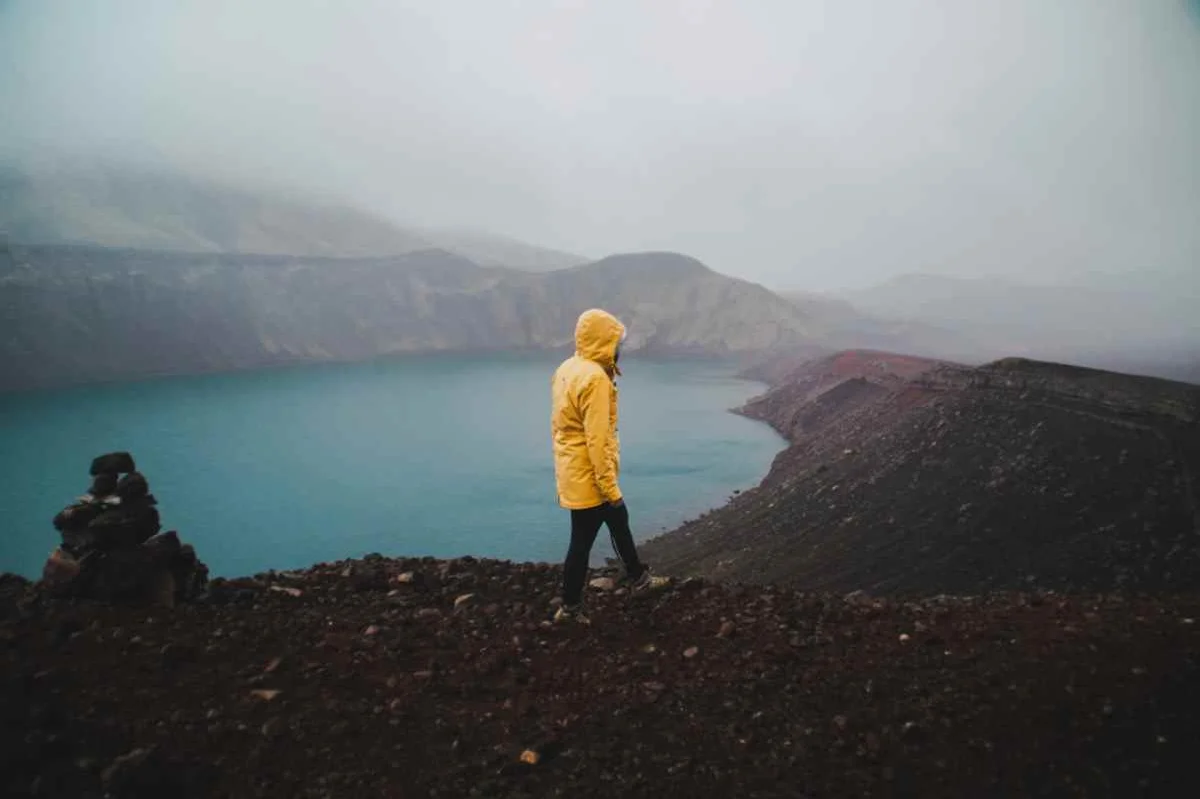 Person in a yellow raincoat standing on a rocky cliff overlooking a misty, blue lake surrounded by mountains in Iceland, illustrating the rugged beauty and changing weather of the region.