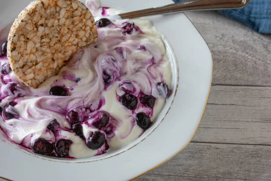 A bowl of creamy yogurt swirled with blueberry compote, topped with a crisp rice cake, placed on a wooden table.