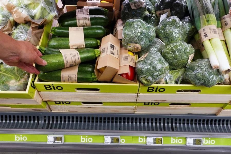 A display of organic vegetables in a supermarket, including zucchinis, broccoli, leeks, and bell peppers, with a hand selecting produce.