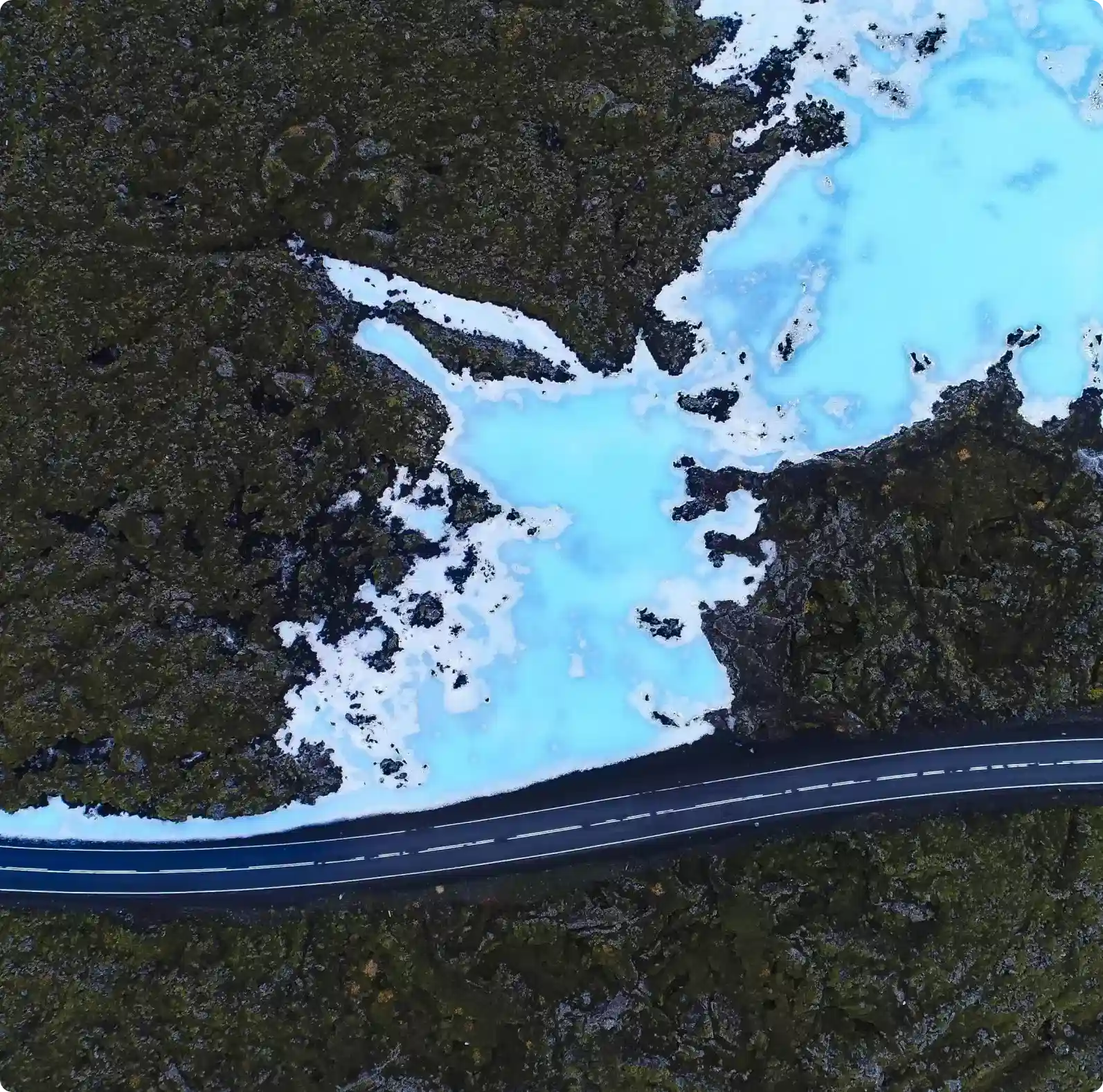 Aerial view of a road cutting through dark lava fields with vibrant blue geothermal water nearby, showcasing the unique landscapes accessible from Reykjavik with a rental car.