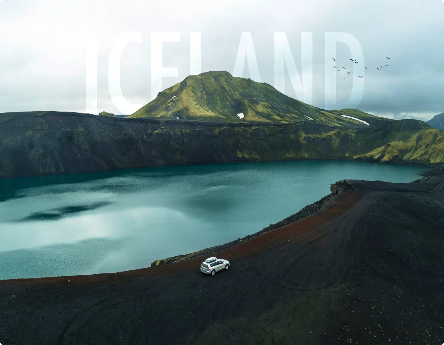 Scenic view of a car parked near a crater lake surrounded by rugged mountains in Iceland, with large 'ICELAND' text overlaying the sky.