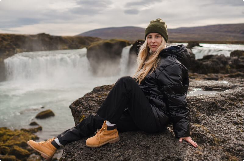 Reisende in Winterkleidung genießen einen atemberaubenden isländischen Sonnenuntergang über einer verschneiten Landschaft mit einem Wasserfall und fernen Bergen.