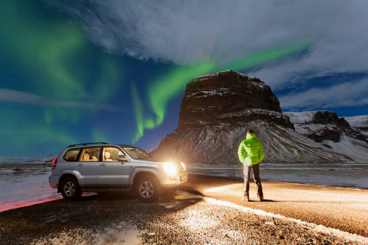 Hombre de pie junto a su coche de alquiler frente a una montaña cubierta de nieve y bajo las luces del norte