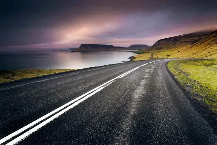 Une autoroute islandaise côtière au crépuscule, avec un ciel dramatique aux teintes violettes et roses se reflétant sur des eaux calmes.