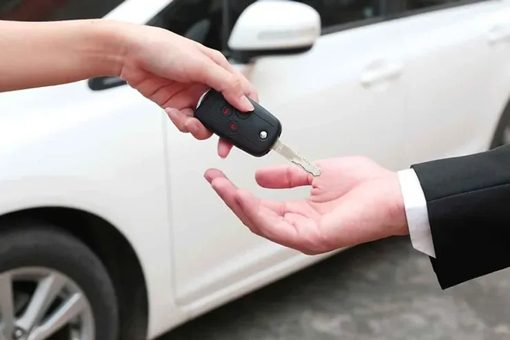 Une personne remettant une clé de voiture à une autre, avec une voiture de location blanche en arrière-plan.