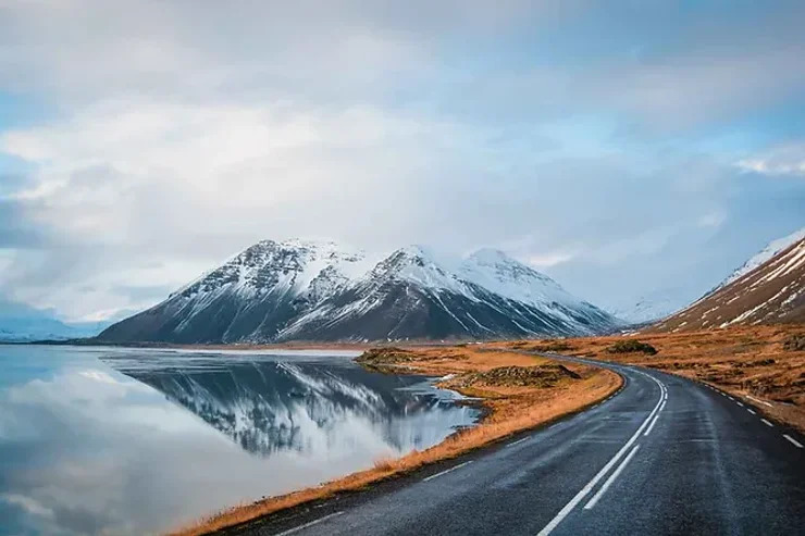 Une route pavée bordée par un lac calme avec des montagnes enneigées reflétées dans l'eau sous un ciel pastel.