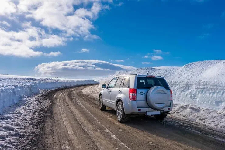 Un SUV blanc sur une route dégagée de neige, entourée de hauts bancs de neige sous un ciel bleu vif en Islande.