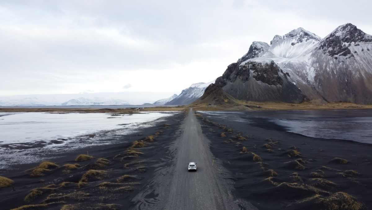 Une voiture blanche roule sur une route de sable noir et de végétation en Islande.