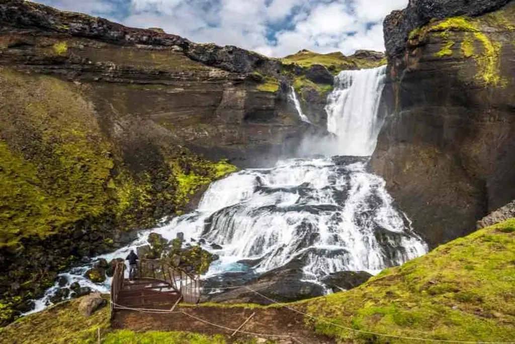 Una stupefacente cascata che scende dalle scogliere di Eldgjá, Islanda, con una piattaforma di osservazione in primo piano, che mostra la bellezza naturale e lo scenario drammatico.