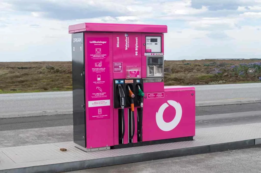 Pompa della stazione di servizio Orkan in Islanda, di colore rosa acceso, inserita in un paesaggio rurale che evidenzia il contrasto tra il colore vivace e l'ambiente naturale circostante.